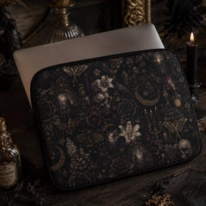 May include: A rectangular laptop sleeve featuring a dark, intricate pattern of moths, crescent moons, skulls, lilies, and candelabras in shades of black, gold, and muted white. The sleeve has a black zippered border and a structured, flat design.