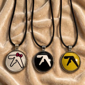 Peut inclure: Trois pendentifs circulaires sur des colliers en cordon noir. Le pendentif de gauche présente un logo Aphex Twin blanc avec un nœud rose. Le pendentif central a un logo blanc sur fond noir. Le pendentif de droite affiche un logo noir sur fond jaune.