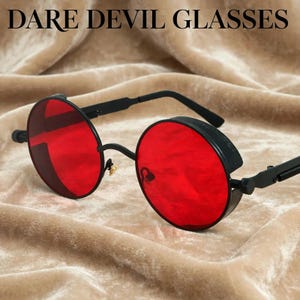 May include: Round sunglasses with vibrant red lenses and a matte black frame. The glasses feature side shields and thin black temples. The text DARE DEVIL GLASSES is printed in black at the top of the frame. The design includes a bridge and nose pads.
