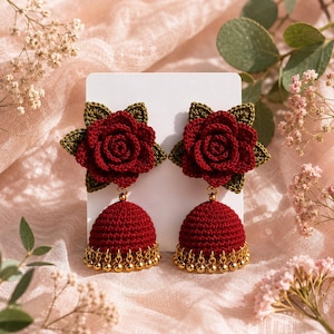May include: A pair of dangle earrings featuring deep red crocheted roses with olive green leaves attached to dark red crocheted jhumka-style domes. The bottom of each dome is adorned with a row of small, shiny gold-toned metallic beads.