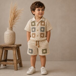 May include: A two-piece crochet outfit consisting of a short-sleeved button-down shirt and matching shorts. The set is made of cream-colored yarn featuring square granny motifs in muted shades of sage green, dusty blue, and tan brown throughout.