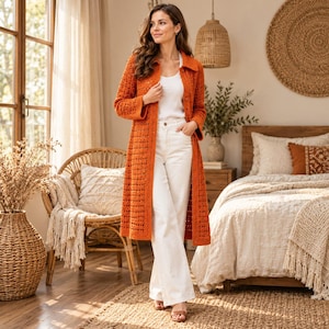 May include: A long, burnt orange open-front cardigan featuring a textured crochet knit pattern with long sleeves, worn over a simple white tank top and white wide-leg trousers. The outfit is completed with brown open-toe sandals.