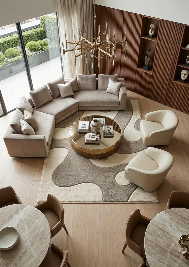 Pu&ograve; includere: Un soggiorno con un grande divano angolare beige, due poltrone color crema e un tavolino rotondo con piano a specchio. Un tappeto fantasia con forme organiche beige e marroni poggia sul pavimento in legno chiaro sotto un lampadario.