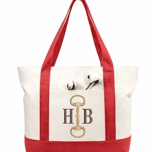 May include: A cream-colored canvas tote bag with red trim and handles. The front features an embroidered design of a gold-colored horse bit centered between the letters H and B in a dark brown serif font. Two cotton bolls sit in the front pocket.