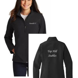 Op de afbeelding: Een zwarte softshell jas met een hoge kraag en een ritssluiting over de gehele lengte. Op de linkerborst is de naam Danielle T. in het wit geborduurd, terwijl op de achterkant de witte geborduurde tekst Top Hill Stables staat.