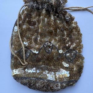May include: A drawstring pouch covered in shimmering bronze and gold sequins. The bag features a decorative zigzag pattern outlined with dark grey seed beads and a horizontal band of lighter, reflective sequins near the bottom edge.