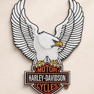 May include: An embroidered patch featuring a white eagle with spread wings perched atop a black and orange Harley-Davidson Motor Cycles logo. The eagle has a yellow beak and black eye details, while the shield-shaped emblem includes white text.