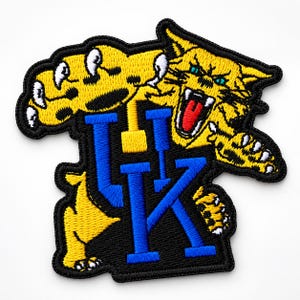 May include: An embroidered patch featuring a yellow wildcat with a red mouth and white teeth, positioned above and behind large blue block letters UK. The design is outlined in black thread, creating a bold, high-contrast sports-themed emblem.