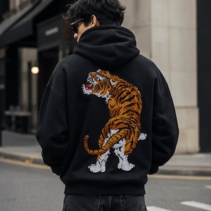 Peut inclure: Un sweat-shirt à capuche noir avec une grande broderie détaillée d'un tigre au dos. Le tigre est représenté avec du fil orange, noir et blanc, dans une pose dynamique, la gueule ouverte, mettant en valeur des coutures et une texture précises.
