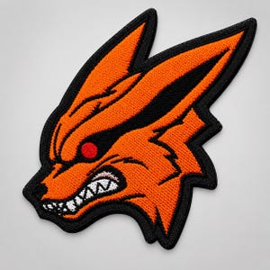 Puede incluir: Un parche bordado que presenta una cabeza de zorro naranja estilizada con orejas puntiagudas, un borde negro y una expresión de gruñido. El diseño incluye un ojo circular rojo brillante, dientes blancos afilados y detalles en negro.