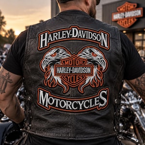 Könnte beinhalten: Eine schwarze Lederweste mit einem großen gestickten Rückenaufnäher mit dem Text Harley-Davidson Motorcycles. Das Design zeigt zwei Adlerköpfe, die ein zentrales Harley-Davidson Motor Cycles Schild-Logo in Orange, Weiß und Schwarz flankieren.