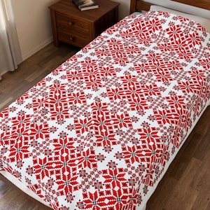 May include: A white bedspread featuring a repeating geometric pattern of red and black snowflakes. The textile covers the surface of a bed, displaying a symmetrical arrangement of star-like shapes across the entire fabric surface.
