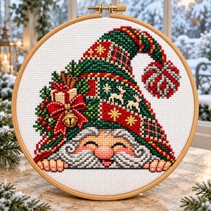 Puede incluir: Un bordado de punto de cruz en un bastidor de madera redondo que muestra un gnomo con una larga barba gris y un gorro grande y flexible. El gorro es verde y rojo con patrones de renos, estrellas y cuadros, decorado con un regalo, un lazo y un cascabel.