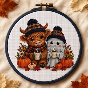May include: A cross-stitch embroidery in a dark circular hoop featuring a brown Highland cow and a white ghost wearing matching plaid hats and scarves. Both figures hold small mugs with steam, surrounded by orange pumpkins and autumn leaves.