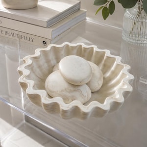 May include: A round, scalloped-edge bowl crafted from cream-colored stone with subtle beige veining. Inside the bowl are three smooth, rounded stones that match the color and texture of the bowl, creating a cohesive and monochromatic appearance.