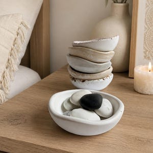 May include: A set of small, shallow, hand-formed ceramic bowls in neutral tones of white, beige, and gray. One bowl is placed in the foreground filled with smooth, rounded river stones in black, white, and gray, while others are stacked behind it.