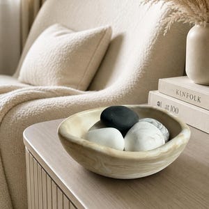 May include: A shallow, round bowl with a beige and white marbled pattern containing several smooth, rounded stones in shades of solid black, solid white, and white with grey veining, resting on a light-toned wooden surface.