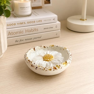 May include: A round, white, speckled trinket dish featuring a sculpted white daisy with a yellow center at its base. The dish has a terrazzo-style pattern with brown and gold flecks along the rim, providing a decorative surface for small items.