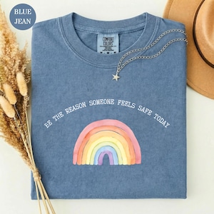 Puede incluir: Una camiseta azul de manga corta con cuello redondo que presenta un gráfico de arcoíris estilo acuarela y el texto blanco "BE THE REASON SOMEONE FEELS SAFE TODAY" impreso sobre el arcoíris. La camiseta tiene una etiqueta de marca visible.