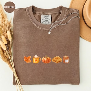 Puede incluir: Una camiseta de cuello redondo marrón con una etiqueta Comfort Colors en el cuello. La parte delantera presenta una fila gráfica de una hoja de otoño naranja, una taza amarilla con un corazón, una calabaza naranja, un suéter beige doblado y una vela naranja encendida en un frasco de vidrio.