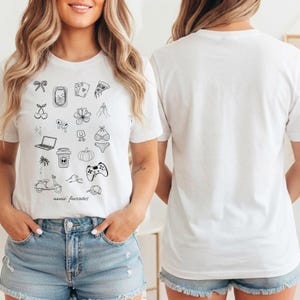 Puede incluir: Una camiseta blanca de manga corta con una colección de iconos de arte lineal negro que incluyen un lazo, cartas de juego, una porción de pizza, cerezas, una flor, un portátil, una taza de café, una palmera, un scooter, un bikini, una calabaza, un mando de juego y el texto "annie' favorites".