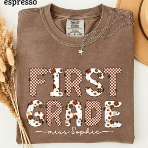 May include: An espresso-colored crewneck t-shirt featuring the text FIRST GRADE in a brown and white checkered pattern with floral accents. Below the text, the name miss Sophie is written in white cursive script. The shirt has a Comfort Colors label.