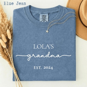 Könnte beinhalten: Ein blaues T-Shirt in Jeansoptik mit weißem Aufdruck LOLA'S grandma EST. 2024. Das Shirt hat einen Rundhalsausschnitt und ein Comfort Colors Etikett am Innenkragen. Eine silberne Halskette mit Sternanhänger liegt auf dem oberen Stoffbereich.