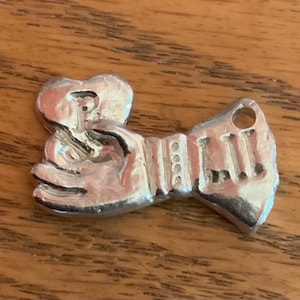 May include: A silver-colored charm in the shape of an accordion. The charm has a heart-shaped top and a rectangular bottom with vertical lines. It has a small hole at the top for attaching to a necklace or bracelet.
