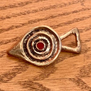 May include: A silver-colored bottle opener with a unique design. The opener features a circular design with concentric rings and a red center. It has a triangular handle. The opener is on a wooden surface.