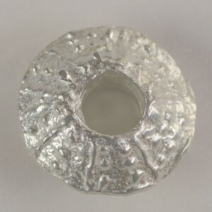 May include: A silver-colored metal bead with a textured surface and a large hole in the center.