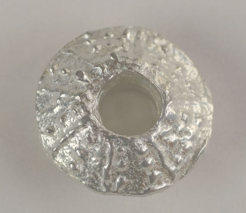 May include: A silver-colored metal bead with a textured surface and a large hole in the center.