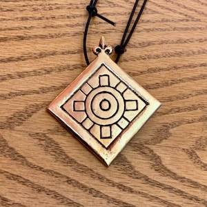 May include: A bronze-coloured pendant necklace with a geometric design. The pendant is diamond-shaped with a sun-like pattern in the centre and a decorative top. The necklace has a black cord.