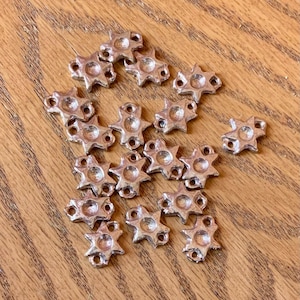 May include: A group of silver-toned metal star-shaped charms with three holes each. The charms are scattered on a wooden surface.