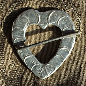May include: A silver heart-shaped brooch with a decorative design and a pin attached.