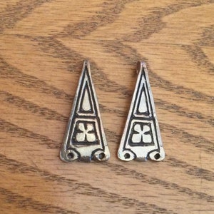 Peut inclure: Deux pendentifs triangulaires en argent avec un trèfle à quatre feuilles au centre. Les pendentifs ont une bordure décorative et trois petites boucles en bas.