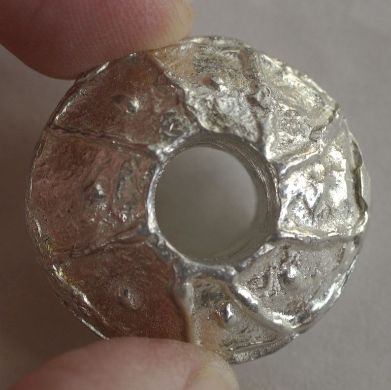 May include: A silver metal bead with a textured surface and a hole in the center. The bead is round and has a slightly irregular shape.