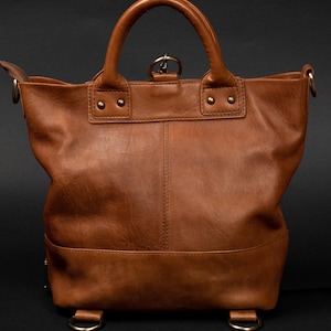 May include: A tan leather tote bag featuring two top handles, a central vertical seam, and brass-toned metal hardware including rivets, side rings, and a top clasp. The bag has a structured base with two lower rings for strap attachments.