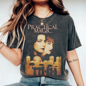 Può includere: Una t-shirt a maniche corte grigio antracite con una stampa grafica di due volti femminili sopra una fila di candele accese e sagome di figure che tengono ombrelli. Il testo "PRACTICAL MAGIC" è stampato in un carattere serif sopra i volti.