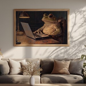 Op de afbeelding: Een ingelijste muurkunstprint met een grote groene kikker die op een donkerhouten stoel aan een bureau zit en op een zilveren laptop typt. Een warme bureaulamp verlicht de scène in een studeerkamer.