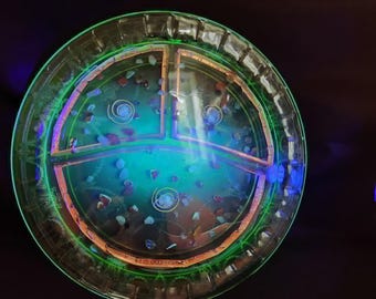 Puede incluir: Una bandeja circular de vidrio verde translúcido con un interior segmentado. La bandeja presenta tres compartimentos distintos que contienen pequeños fragmentos de piedra multicolores y espirales metálicas, todo brillando bajo luz ultravioleta.