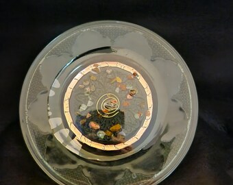 Puede incluir: Un plato circular de vidrio con una incrustación central de resina. La incrustación está bordeada por un anillo de color cobre y contiene pequeños fragmentos de piedra multicolores y un alambre en espiral de metal dorado.