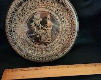 Puede incluir: Un plato decorativo redondo con intrincadas figuras de estilo egipcio en tonos dorados y patrones inspirados en jeroglíficos sobre un fondo oscuro. Una regla de madera colocada debajo muestra un diámetro de unos 25,4 cm en la superficie.