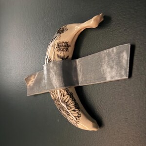 May include: A cream-colored banana sculpture featuring detailed black ink illustrations of sunflowers and botanical elements, secured to a dark surface with a single horizontal strip of metallic silver duct tape across its center.