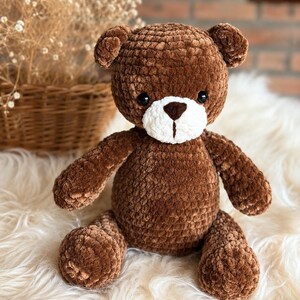 May include: A brown crocheted teddy bear with a soft, plush texture. The bear features a white muzzle, a dark brown stitched nose, and small black bead eyes. It is crafted from thick, velvety yarn and has rounded ears, arms, and legs.