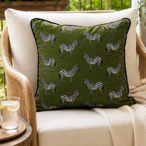 May include: A square throw pillow featuring an olive green fabric with a repeating pattern of black and white zebras. The pillow has black piping along the edges and is positioned on a light-colored seat cushion with a cream-colored throw blanket.