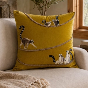 May include: A square mustard yellow throw pillow featuring a series of embroidered ring-tailed lemurs. The lemurs are depicted in various poses across the surface, separated by three horizontal, wavy lines of textured, light brown jute-like trim.