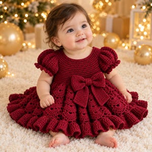 May include: A deep burgundy crochet dress featuring short puffed sleeves, a round neckline, and a large central bow at the waist. The skirt is designed with a textured scalloped pattern and a ruffled hemline, crafted from yarn.