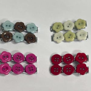 May include: Four pairs of hair clips, each featuring three flower-shaped buttons attached to a metal bar. The sets include light blue and brown, pale yellow and cream, bright pink, and deep red colour combinations with visible button holes.