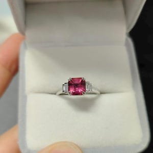 Könnte beinhalten: Ein silberfarbener Ring mit einem zentralen, smaragdgeschliffenen rosa Edelstein, der auf jeder Seite von zwei kleineren, im Baguetteschliff geschliffenen klaren Steinen flankiert wird, präsentiert in einer hellgrauen, samtgefütterten Schmuckschatulle.