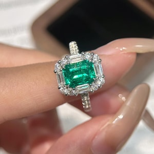Könnte beinhalten: Ein silberfarbener Ring mit einem rechteckigen, smaragdgeschliffenen grünen Edelstein in der Mitte, umgeben von einem Kranz aus kleinen runden Diamanten und Baguette-Diamanten, auf einer Ringschiene mit kleinen runden Diamanten besetzt.